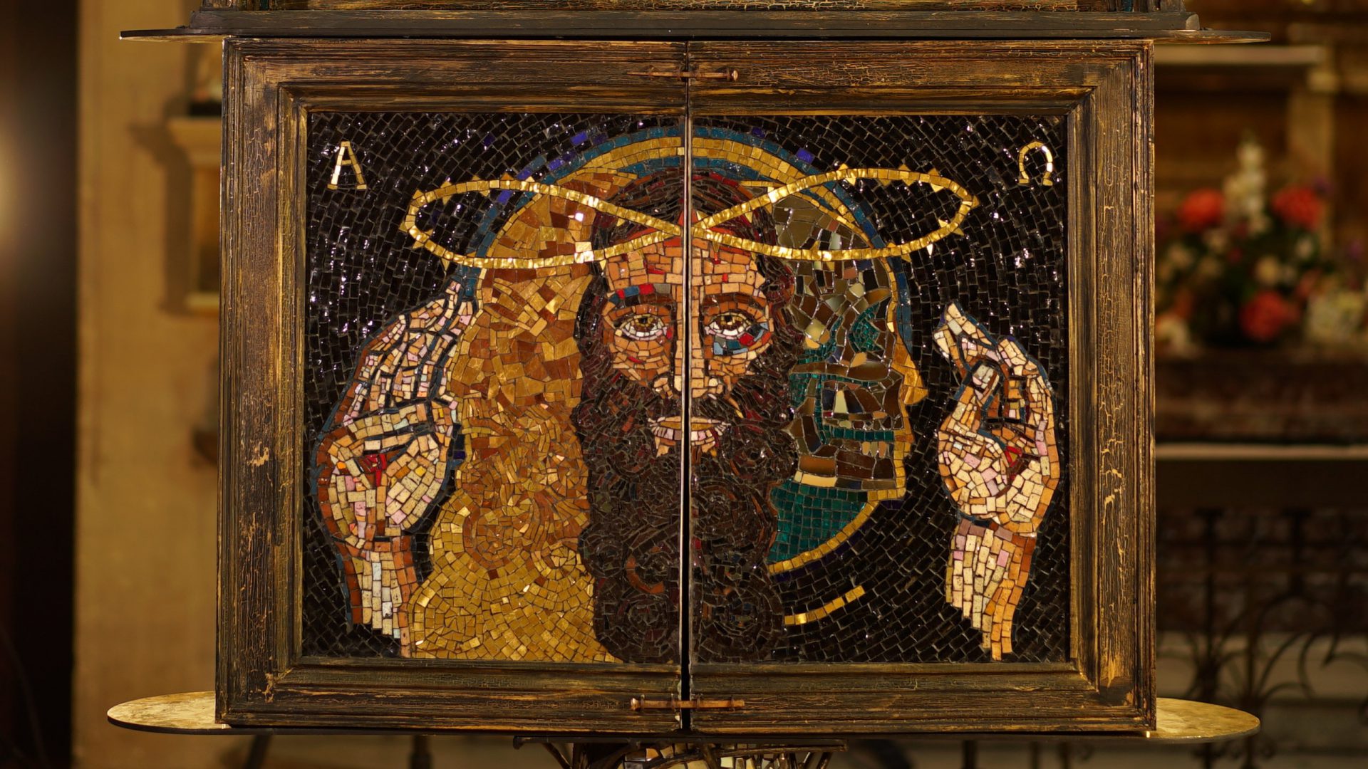 Jesus’ face is in the centre, and the doors open at the point of his mouth and lips so as he speaks, the doors open and vice versa. The doors close using nails reminiscent of his death. Jesus is not blue-eyed and western but dark, battered and bruised from the Passion and his skin reflects his true ethnicity from the Mediterranean. Jesus’ crown of thorns are made from the infinity symbol, for eternity. He is the eternal one with an eternal message, and this symbol also unites science and religion within God’s kingdom. Eternity is echoed too in the use of the gold Alpha and Omega symbols in the top left and right corners – the two Greek letters for the beginning and the end of the alphabet as God is the beginning and the end of all creation. The Holy Spirit appears in a number of forms in this artwork but here on the right within the Trinity first as glass and mirror, to help us understand how the Holy Spirit reflects God and Christ and we find ourselves in God by looking in the mirror of the Holy Spirit. The Holy Spirit is also conveyed here with open hands around the central Trinity but with the stigmata in them to represent the nails in Christ’s hands when crucified. God is depicted as 24 carat gold on the left side – with different types of gold to express his perfect totality as only the best will do for God.